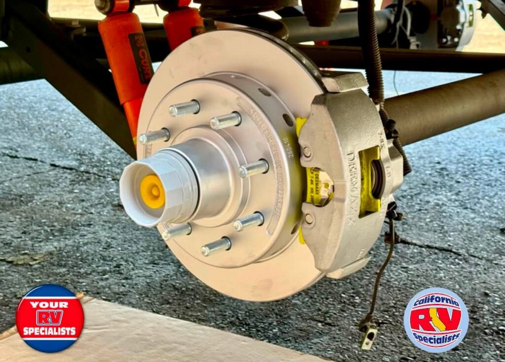 DeeMaxx Disc Brakes - California RV Specialists Close-up of a RV trailer wheel hub with brake rotor and caliper, showing lug studs and a yellow center cap on a paved surface.