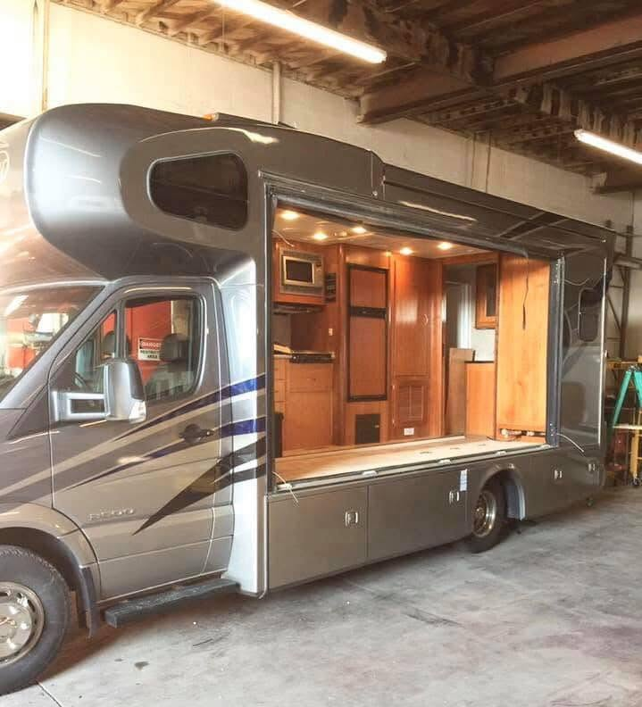 Grey motorhome with its side slide-out extended, revealing a wood-paneled kitchen and living area inside a garage-like space.