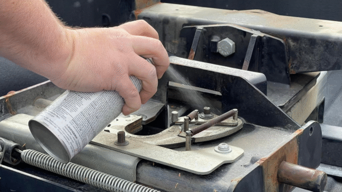 Close-up of a hand spraying lubricant onto a rusty trailer hitch frame.