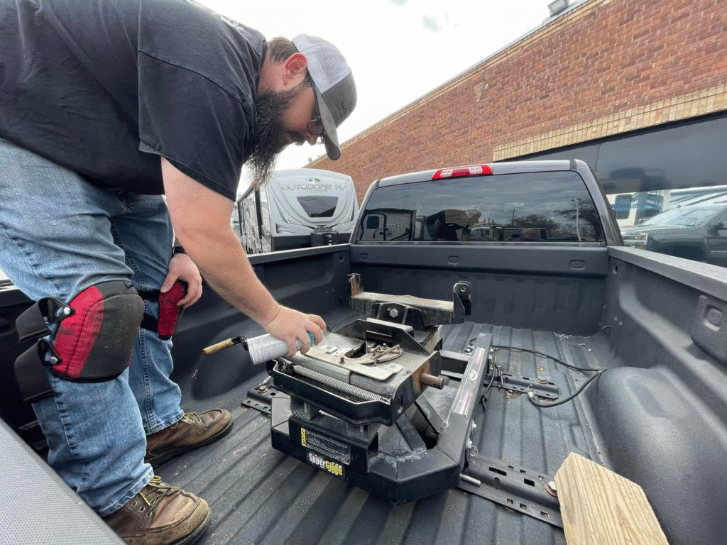 image - California RV Specialists Bearded man in a cap kneels in a pickup bed, using a tool to work on a device inside the toolbox area beside a brick wall outside.