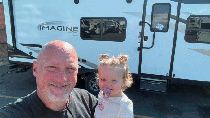 Man and young child pose for a selfie in front of a large white travel trailer with 'IMAGINE' branding in a dealership lot.