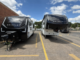 Two black Brinkley fifth-wheel RVs parked side by side in a lot, with a brick wall on the left and a blue sky above.