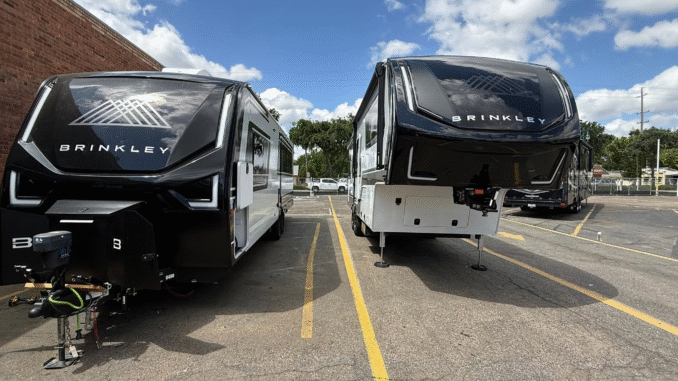 Two black Brinkley fifth-wheel RVs parked side by side in a lot, with a brick wall on the left and a blue sky above.