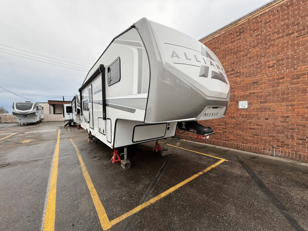Large white fifth-wheel RV parked in a lot beside a red brick building with another RV in the background