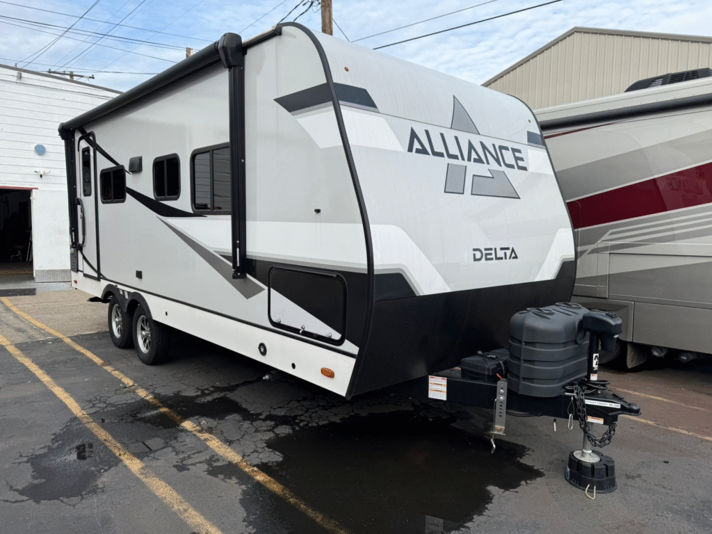 White Alliance Delta travel trailer with gray and black graphics, connected to a hitch in a parking lot, propane tanks in front.