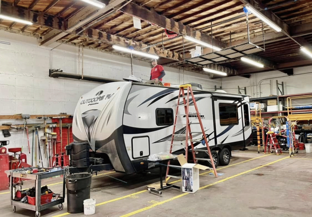 RV trailer inside a workshop being serviced, man on roof with ladders and tools nearby