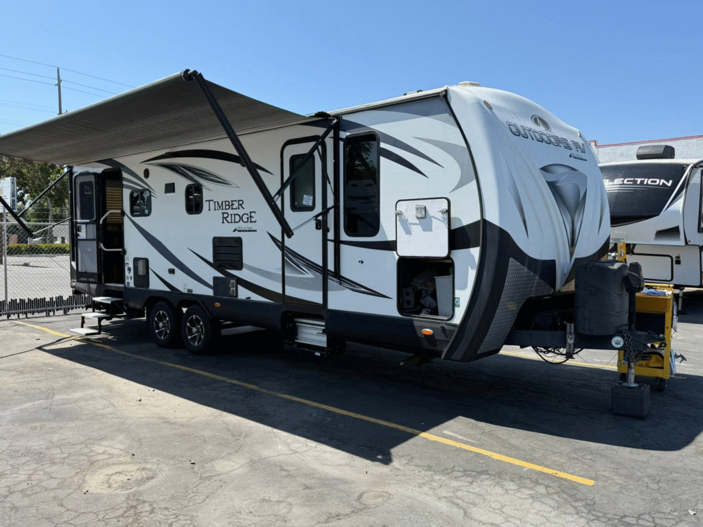 A large white travel trailer with an extended awning, parked in a lot with a chain-link fence in the background and another trailer nearby.