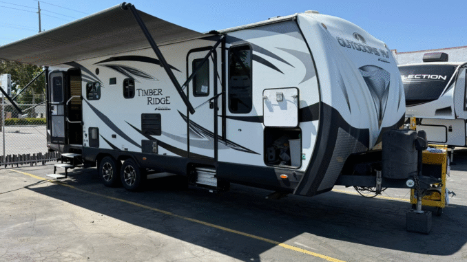 A large white travel trailer with an extended awning, parked in a lot with a chain-link fence in the background and another trailer nearby.