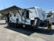 A large white travel trailer with an extended awning, parked in a lot with a chain-link fence in the background and another trailer nearby.