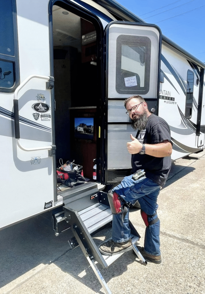 Bearded man in a black T-shirt giving a thumbs up while stepping down from an RV with extendable metal stairs under a blue sky.