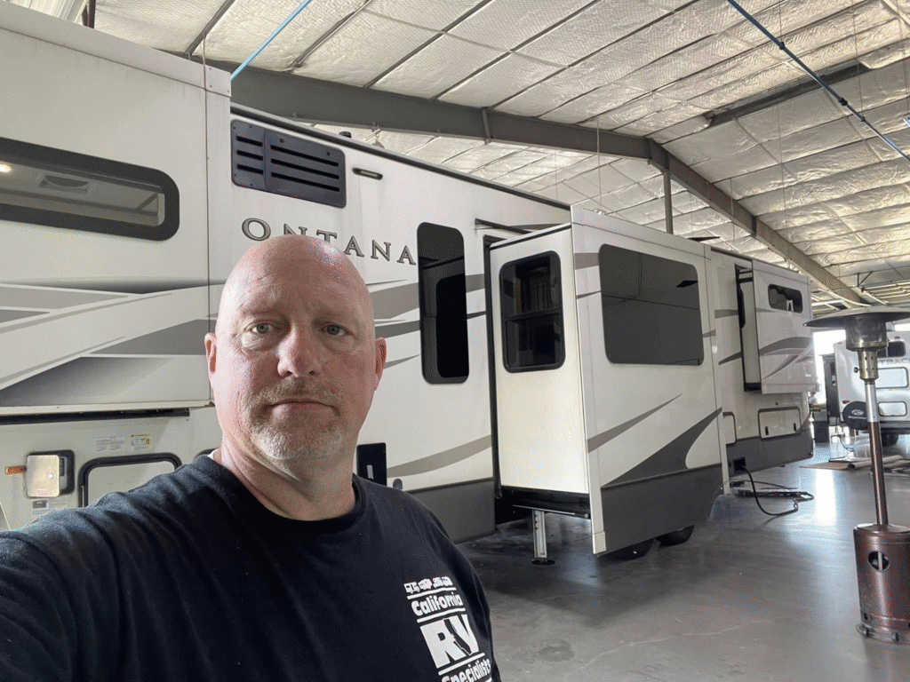 Selfie of a man in a showroom with large white recreational vehicles (RVs) parked in a row behind him inside a spacious, well-lit warehouse-like space.