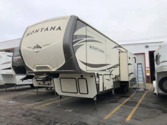 A large Montana fifth-wheel RV parked in a lot, with a ladder and other trailers nearby. The front cap and side graphics are visible, including the word Montana on the front and side panels.