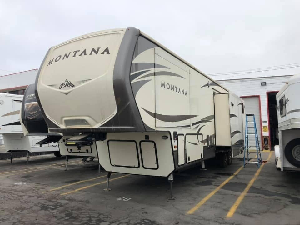 A large Montana fifth-wheel RV parked in a lot, with a ladder and other trailers nearby. The front cap and side graphics are visible, including the word Montana on the front and side panels.