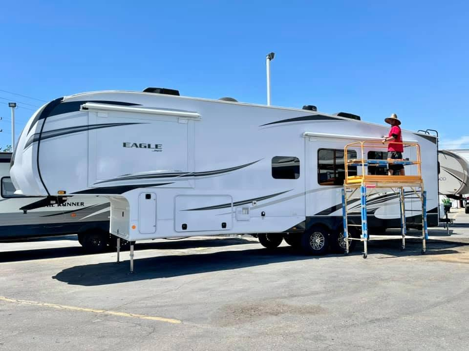 Large white RV trailer with blue accents; a person on a yellow scaffold works near the side door in a sunny lot.