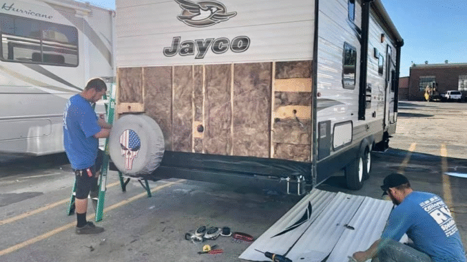 Two workers repair the exterior of a Jayco travel trailer; siding is removed and tools are on the ground.