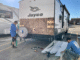 Two workers repair the exterior of a Jayco travel trailer; siding is removed and tools are on the ground.