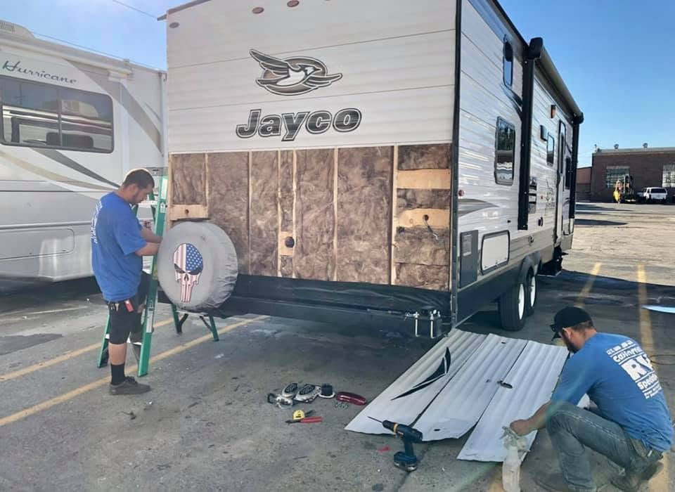 Two workers repair the exterior of a Jayco travel trailer; siding is removed and tools are on the ground.
