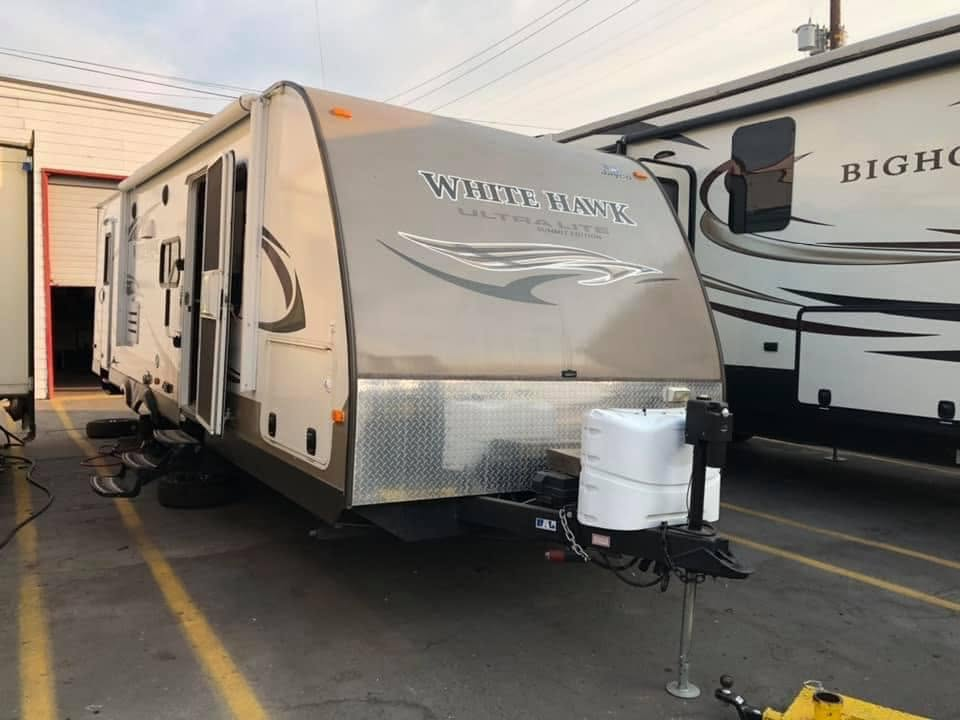 A gray travel trailer labeled 'White Hawk' parked in a lot beside a larger RV, with a diamond-plate front and a propane tank on the hitch.