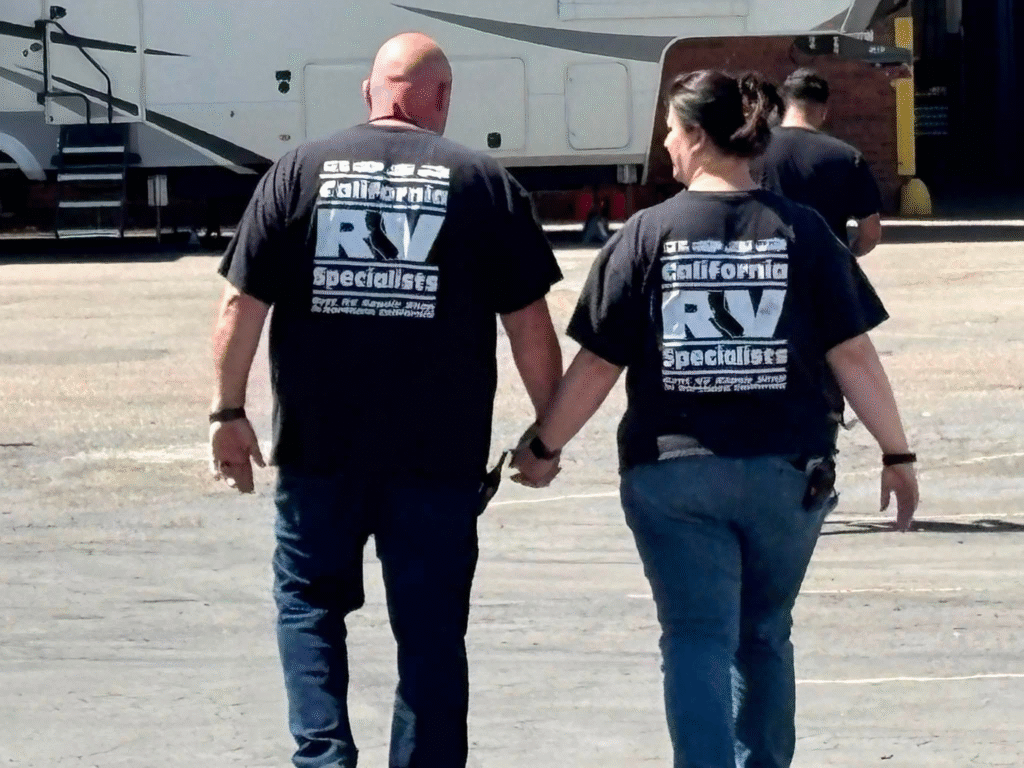 Two adults walk away hand in hand on a tarmac beside a parked recreational vehicle, both wearing black tees with large white text on the back.