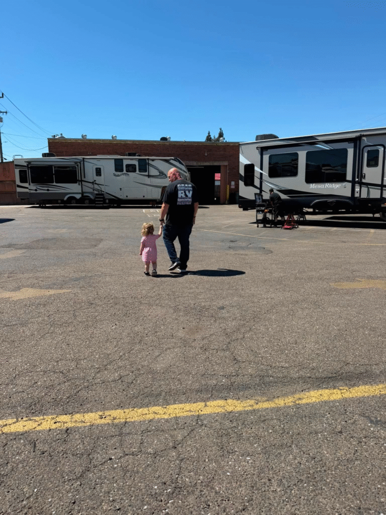 Man and child walk hand in hand past parked motorhomes in a lot under a clear blue sky, with a brick building in the background.