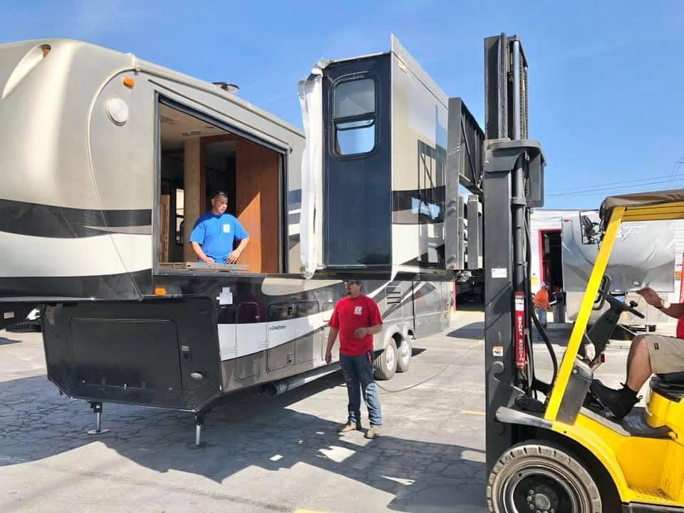 A man in a red shirt watches as a forklift moves a large fifth-wheel trailer into a truck.