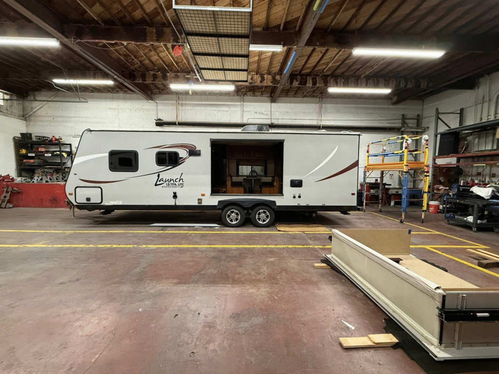 Indoor workshop with a white RV trailer parked inside; open side door and work shelving on the left, scaffolding and equipment on the right, bright overhead lights.