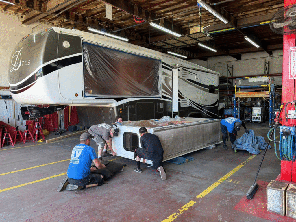 Mechanics kneeling and working on the underside of a large RV inside a workshop, with plastic covering a slide-out panel.