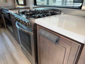 Stainless steel gas range with four burners in a compact kitchen, next to a white countertop and window with blinds.