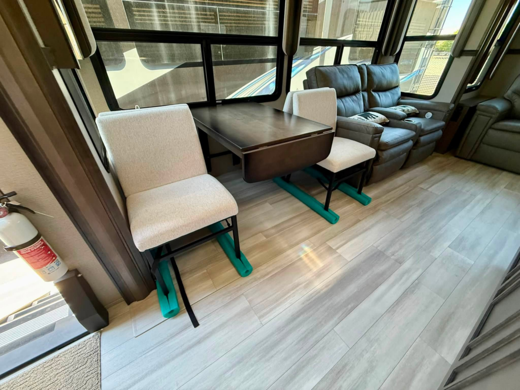 Interior seating area of a vehicle with a dark wood table, beige chairs, and a gray recliner sofa beside large windows; a fire extinguisher is near the door.