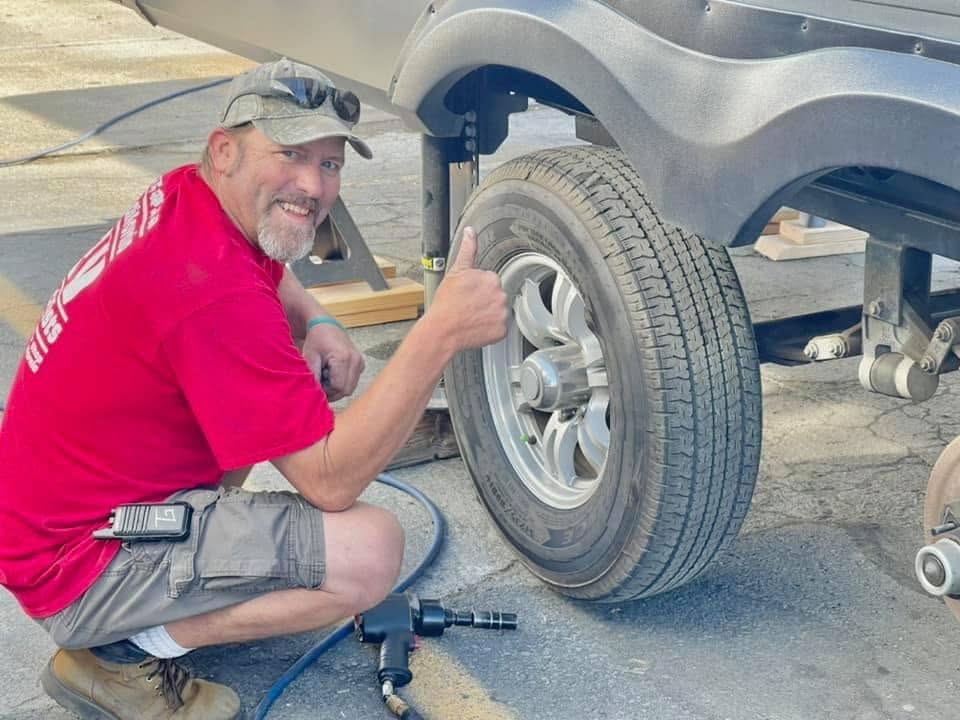 Man kneels beside a trailer wheel on pavement, giving a thumbs-up while using a pneumatic tool on the tire.
