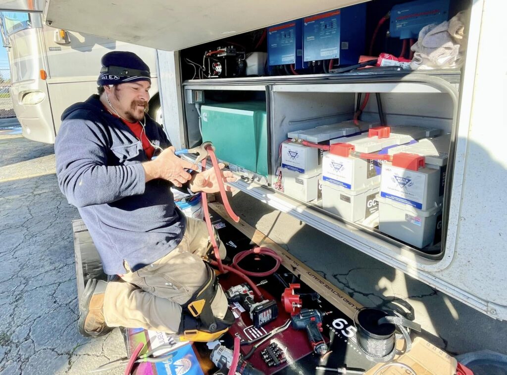 Technician kneeling beside an open service van, handling red jumper cables among batteries and tools inside the compartment.