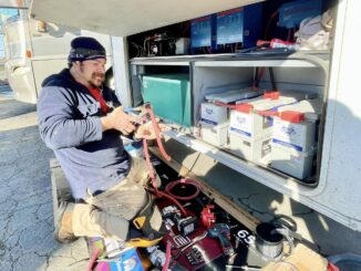 Technician kneeling beside an open service van, handling red jumper cables among batteries and tools inside the compartment.