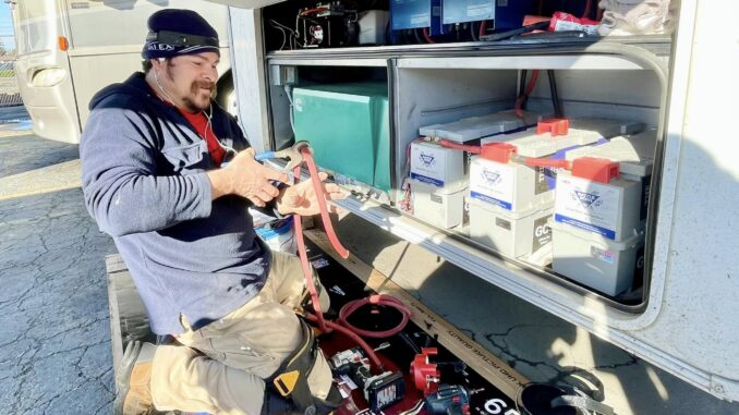Technician kneeling beside an open service van, handling red jumper cables among batteries and tools inside the compartment.