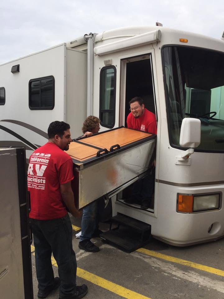 Three workers in red shirts lift a wooden piano into a white RV through its side door, using a ramp and straps for moving.