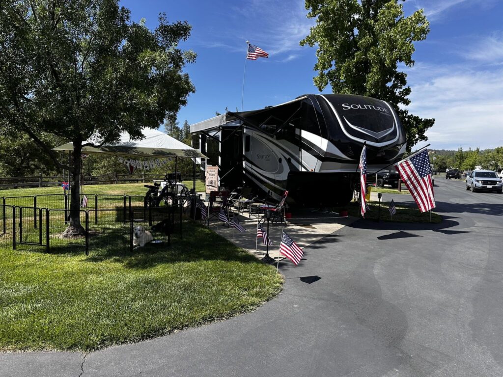 Large black-and-white Solitude RV parked at a campground with American flags, a white canopy, bicycles, and green trees under a blue sky.