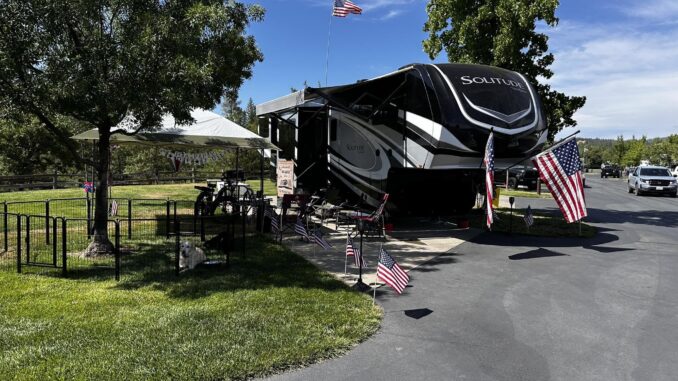 Large black-and-white Solitude RV parked at a campsite with American flags, canopy, and bicycles in a sunny park.