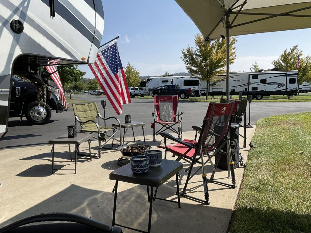 RV park scene with large motorhomes, American flags, and folding chairs set up under a canopy on a sunny day.