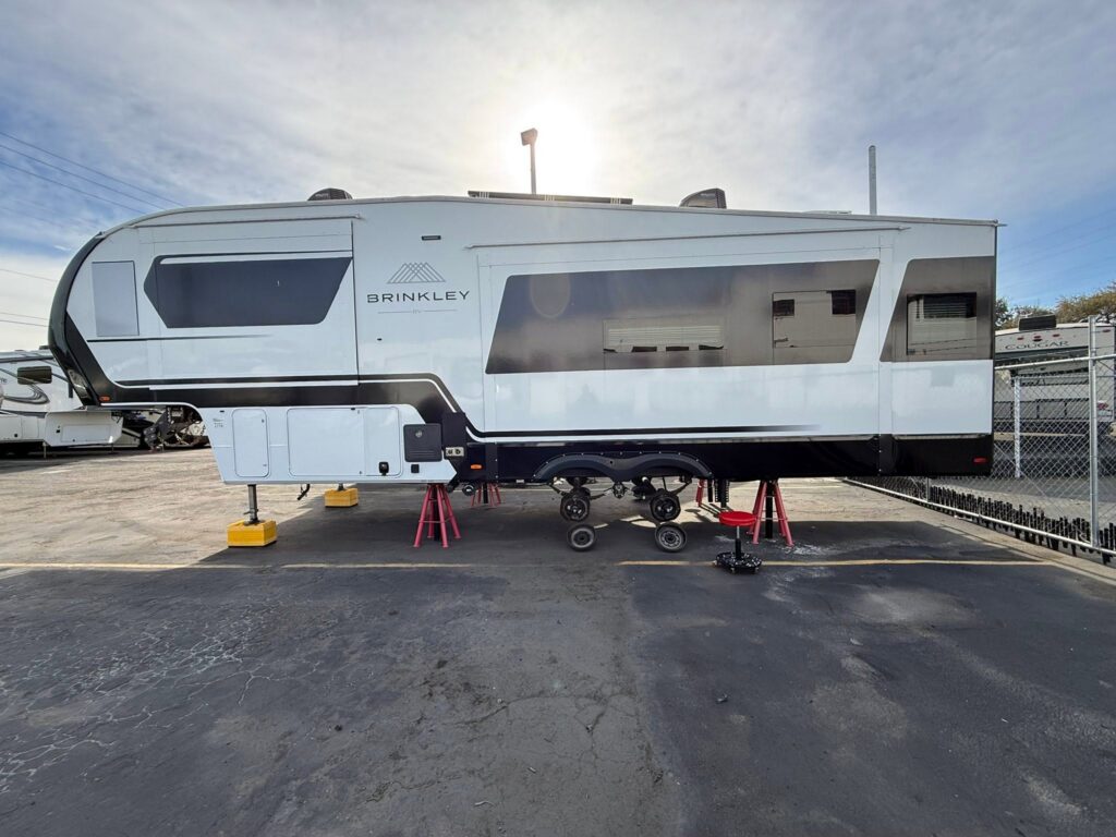Side view of a white Brinkley RV trailer raised on stands in a lot with a chain-link fence and clear sky above