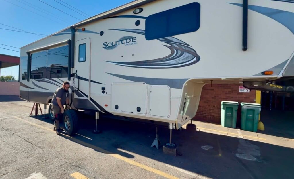 Large Solitude RV being serviced in a parking lot; a man works on the front wheel area.