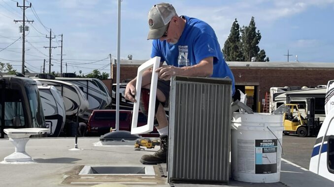 Worker in a blue shirt and gray cap leaning over a white window frame, repairing or installing it in an outdoor RV repair yard.