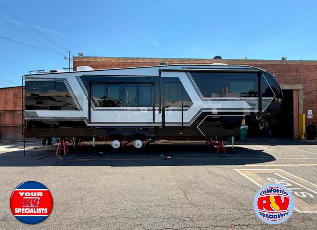 Large modern travel trailer on leveling jacks outside a service shop, with branding logos in the corners: 'Your RV Specialists' and 'California RV Specialists'