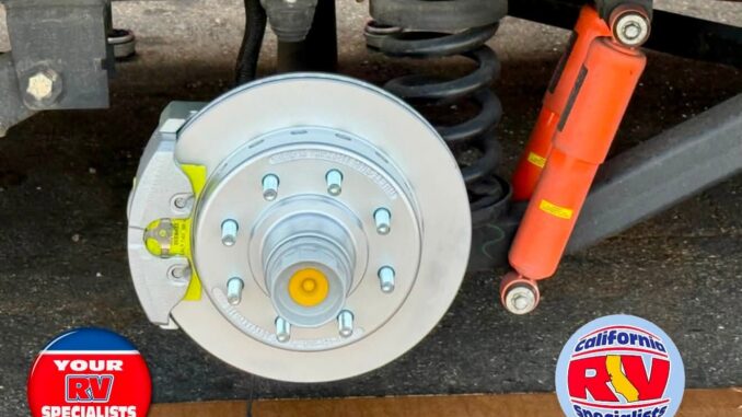 RV trailer undercarriage showing exposed brake rotor, hub, coil spring, and orange shock absorbers; logos read 'Your RV Specialists' and 'California RV Specialists'