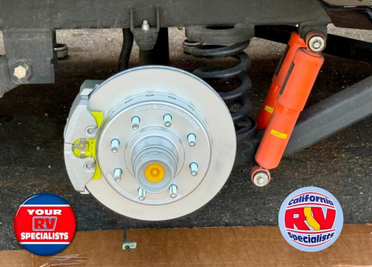RV trailer undercarriage showing exposed brake rotor, hub, coil spring, and orange shock absorbers; logos read 'Your RV Specialists' and 'California RV Specialists'