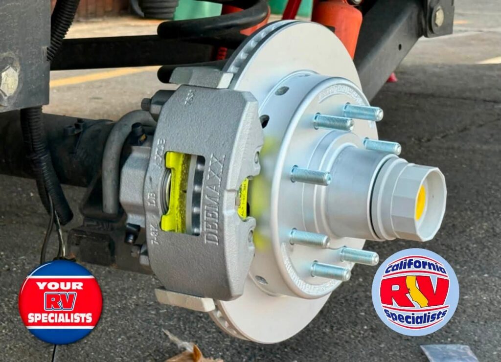 Close-up of a vehicle brake assembly showing a silver rotor, gray brake caliper, and exposed lug studs, with RV specialist logos in the corners.