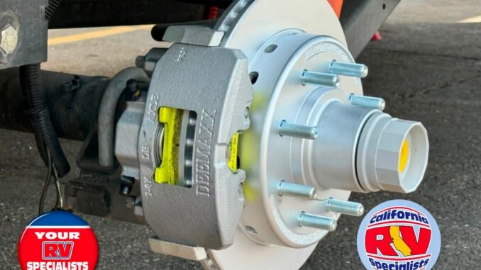 Close-up of a vehicle brake assembly showing a silver rotor, gray brake caliper, and exposed lug studs, with RV specialist logos in the corners.