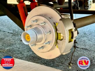 Close-up of a RV trailer wheel hub with brake rotor and caliper, showing lug studs and a yellow center cap on a paved surface.